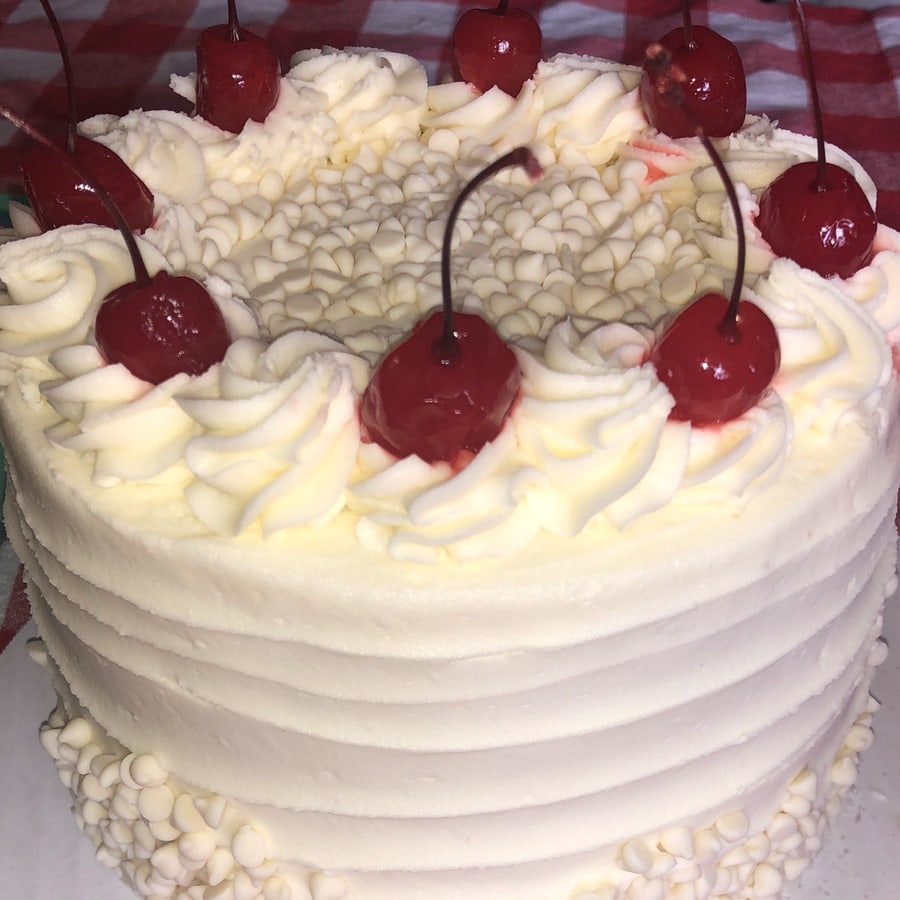 A homemade layer cake in white buttercream, ringed with piped swirls and maraschino cherries.