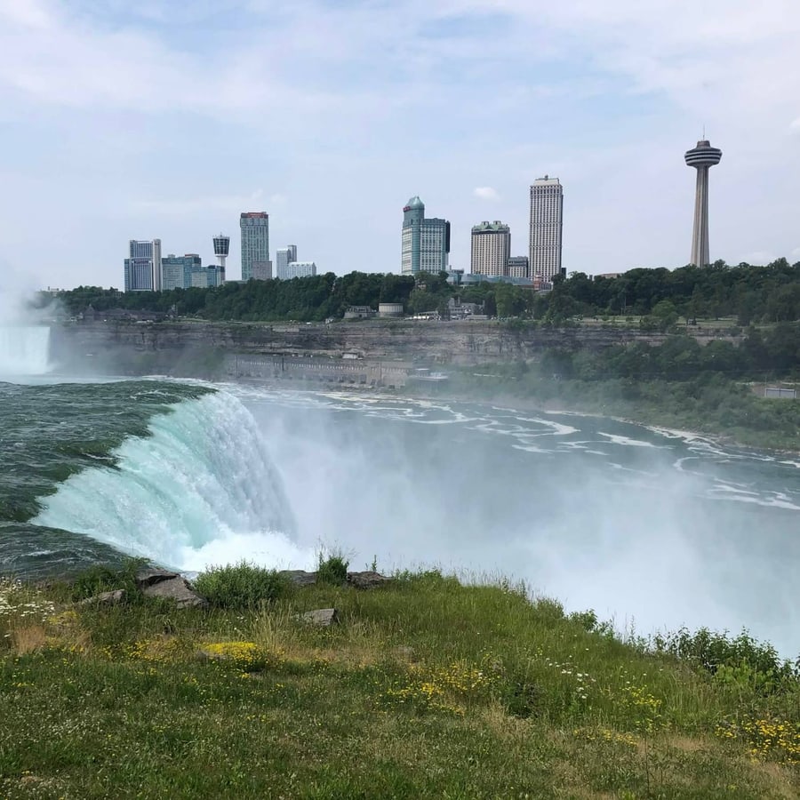 Niagara Falls pouring over the edge in a cloud of mist, with the city skyline and a tall observation tower beyond.